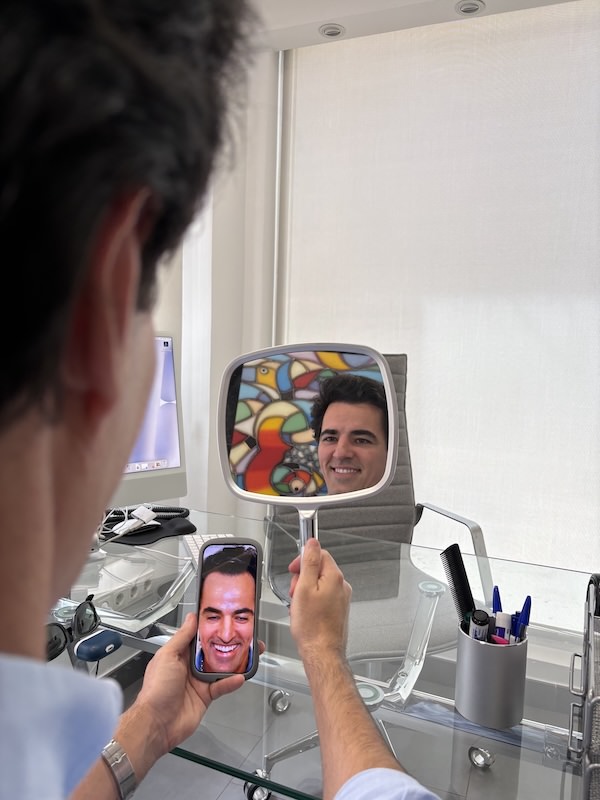 Paciente masculino joven en una consulta médica, mirándose en un espejo de mano y en su smartphone, que muestra una foto de su rostro, disfrutando de su nueva línea capilar y el resultado exitoso de su injerto capilar en Folika Hair Clinic, Madrid.